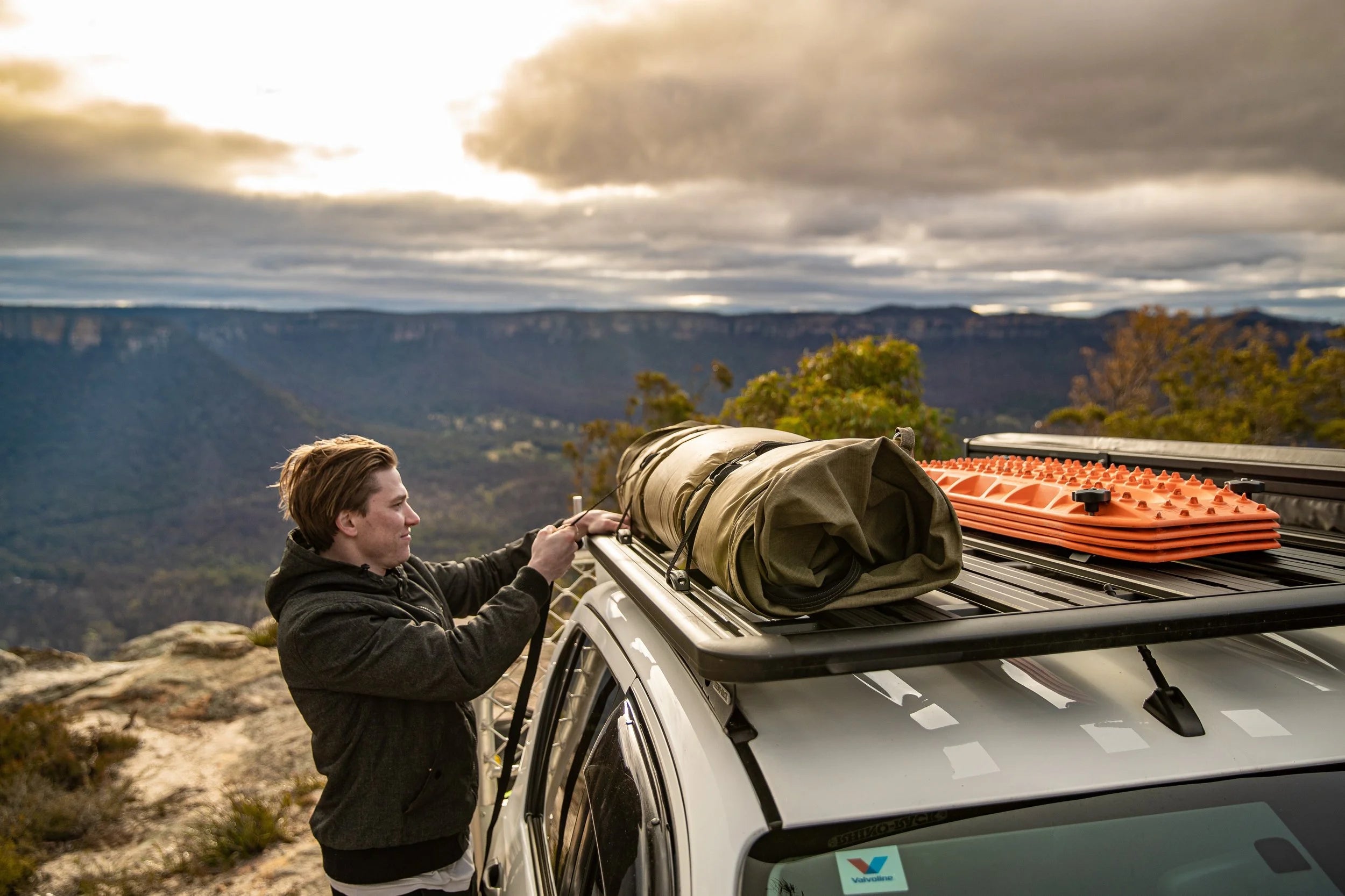 Person securing camping gear on a car roof with a scenic mountain backdrop